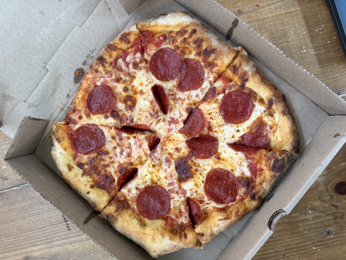 A wood-fired pepperoni pizza with a blistered, charred crust in an open takeout box from Stoney's Fired Pizza